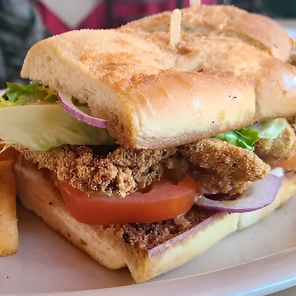 Close-up of a fried fish po'boy sandwich with lettuce, tomato, and red onion on a white plate.
