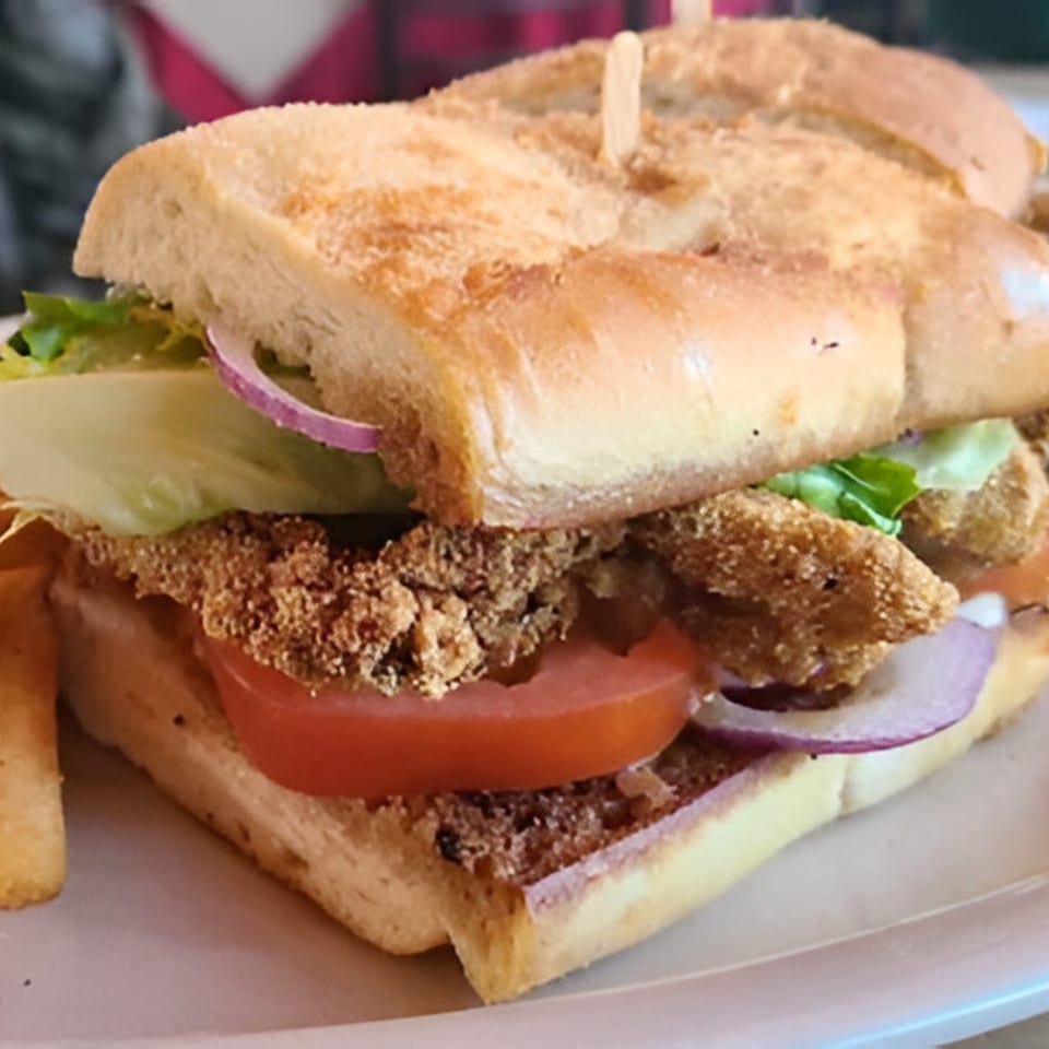 Close-up of a fried fish po'boy sandwich with lettuce, tomato, and red onion on a white plate.