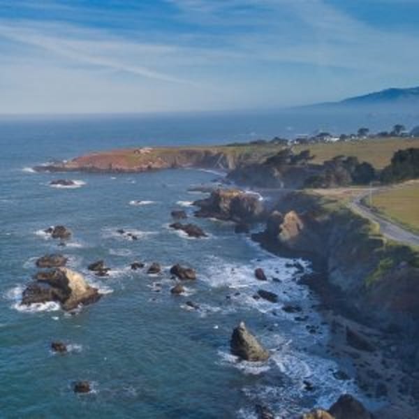 Rugged California coastline with rocky shores, crashing waves, and a winding road along the cliffs.