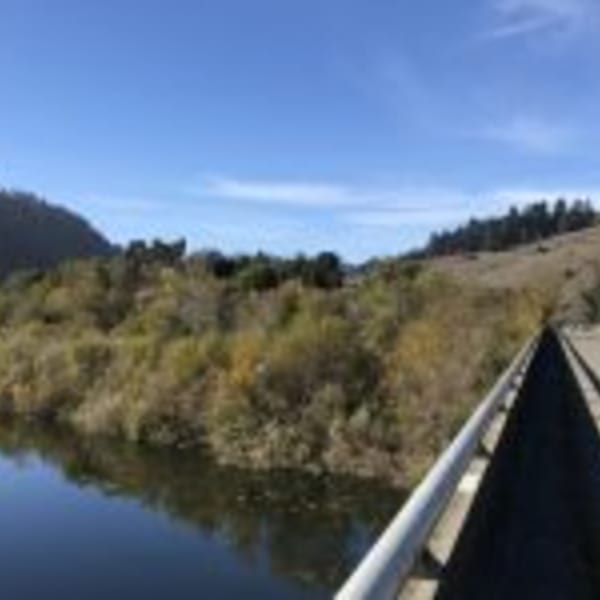 A bridge over a calm body of water, surrounded by lush green and brown hills under a clear blue sky.