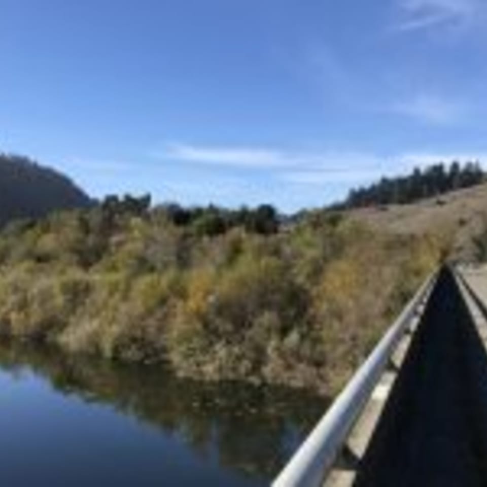 A bridge over a calm body of water, surrounded by lush green and brown hills under a clear blue sky.
