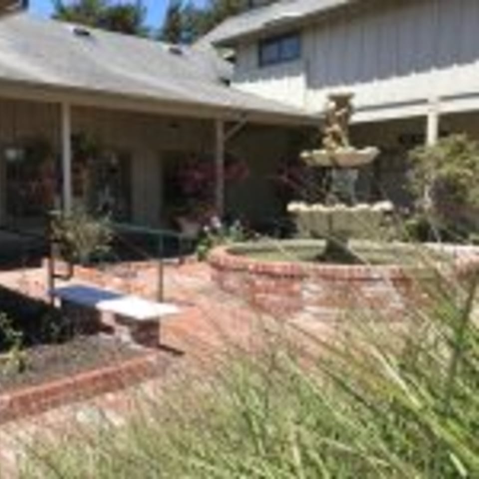 Outdoor patio with a brick fountain, lush greenery, and a building in the background.