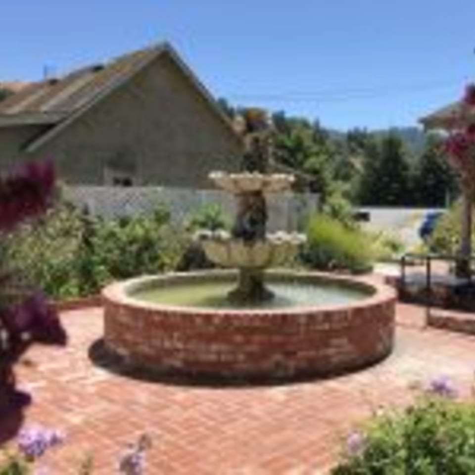 A two-tiered stone fountain with a brick base sits on a brick patio, surrounded by lush greenery.