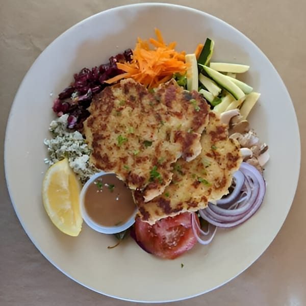 Two crab cakes with a side of shredded carrots, zucchini, red onion, tomato, and a lemon wedge.